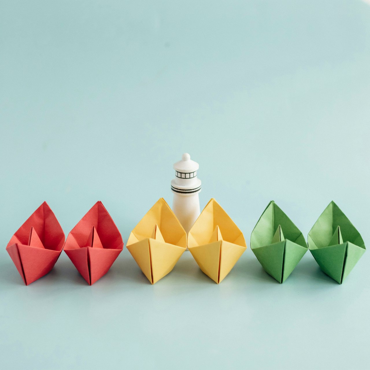 Exactly six folded paper boats sit on a pale blue tabletop: two red boats on the left, two yellow boats in the center, and two green boats on the right. A small white lighthouse figurine stands behind the center pair, nothing else. — Ideogram v4.5