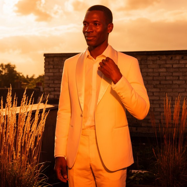 Under the copper glow of a rooftop terrace, an adult Black man in a tailored cream dinner suit is caught smoothing his lapel as evening breeze stirs nearby grasses; calm poise, sculpted shadows and refined fashion-magazine polish. — Krea 2 Large