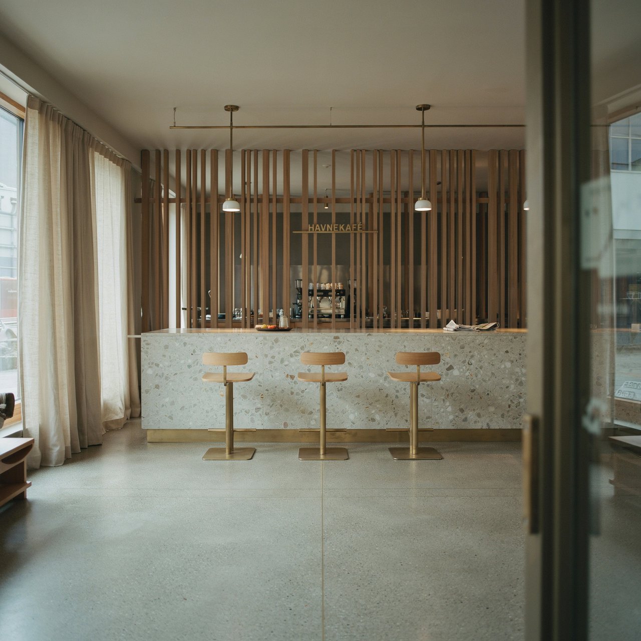 Inside an empty harbor café, restrained Danish modern design pairs ash slat screens, pale terrazzo, and brushed brass; overcast window light reaches the counter, viewed from the entrance, with aligned joinery and believable supports. — Ideogram v4