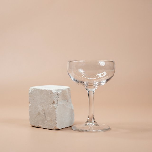 Beside a pale limestone block, an unbranded handblown cocktail coupe in clear soda-lime glass catches crisp window-shaped highlights; its curved bowl bends a muted apricot backdrop under broad diffused key light and narrow cool rim lighting, commercial product image, no lettering. — Ideogram v4