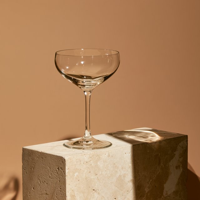 Beside a pale limestone block, an unbranded handblown cocktail coupe in clear soda-lime glass catches crisp window-shaped highlights; its curved bowl bends a muted apricot backdrop under broad diffused key light and narrow cool rim lighting, commercial product image, no lettering. — Midjourney V8.2