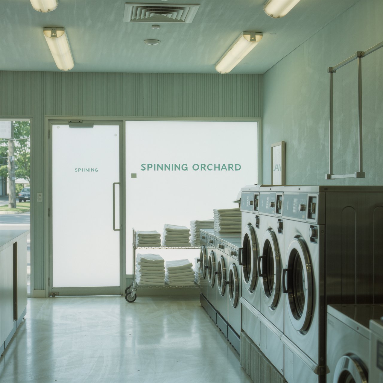 Inside a quiet neighborhood laundromat, chrome machines and folded linen glow beneath cool ceiling lights; "SPINNING ORCHARD" is printed in pale mint letters on the frosted glass entry door. — Kling v3 Omni
