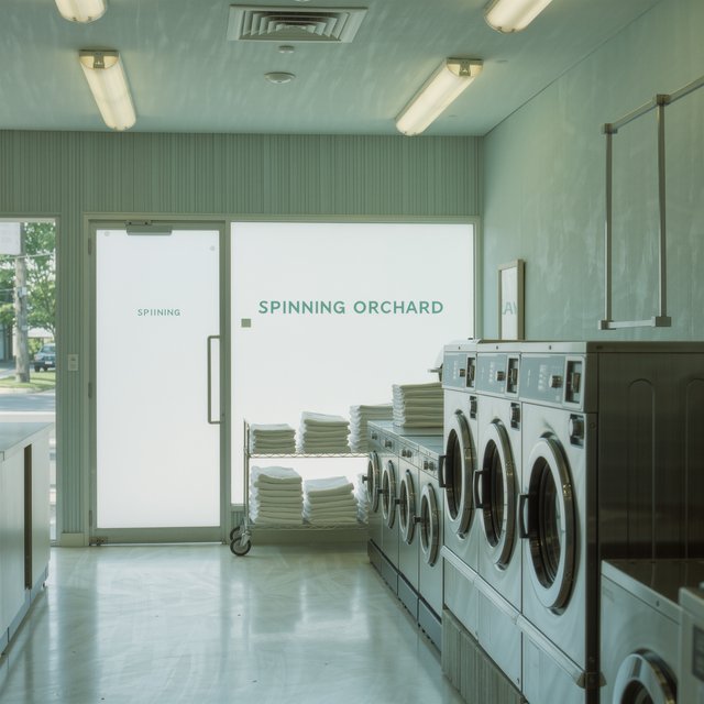 Inside a quiet neighborhood laundromat, chrome machines and folded linen glow beneath cool ceiling lights; "SPINNING ORCHARD" is printed in pale mint letters on the frosted glass entry door. — Kling v3 Omni