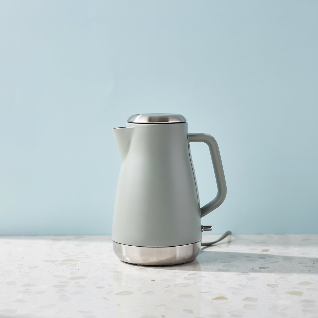 Commercial product photograph of an unbranded electric kettle made from matte sage ceramic and brushed stainless steel, resting on a white terrazzo surface against a pale blue background, with soft window light from the left and a delicate grounding shadow. — Ideogram v4