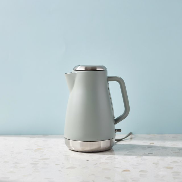 Commercial product photograph of an unbranded electric kettle made from matte sage ceramic and brushed stainless steel, resting on a white terrazzo surface against a pale blue background, with soft window light from the left and a delicate grounding shadow. — Ideogram v4