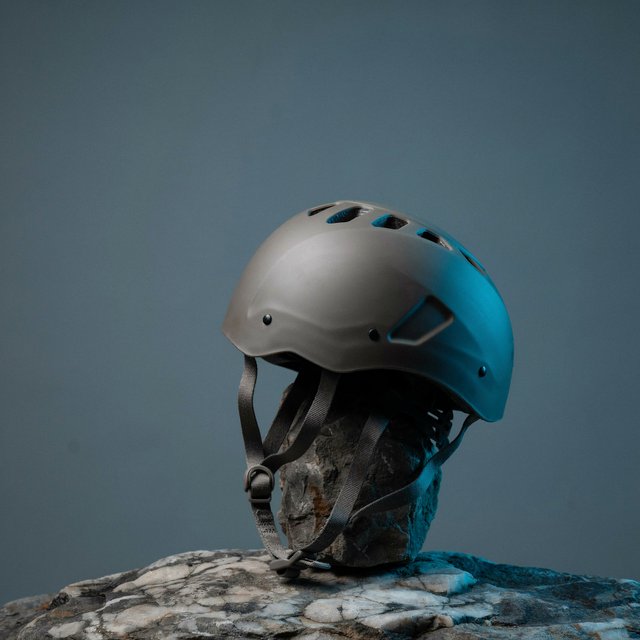 Against a muted slate backdrop, an unbranded climbing helmet with a matte recycled-polymer shell and woven nylon straps rests on rough granite; broad diffused key light and a cool rim light shape this commercial product image, with no lettering. — Ideogram v4.5
