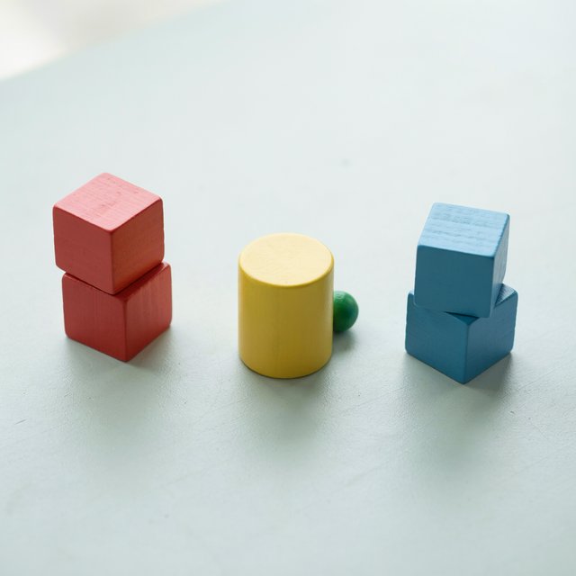 Exactly five wooden toy blocks sit on a pale gray table: two red cubes on the left, one yellow cylinder in the center, and two blue cubes on the right. A small green ball sits behind the cylinder, nothing else. — Ideogram v4