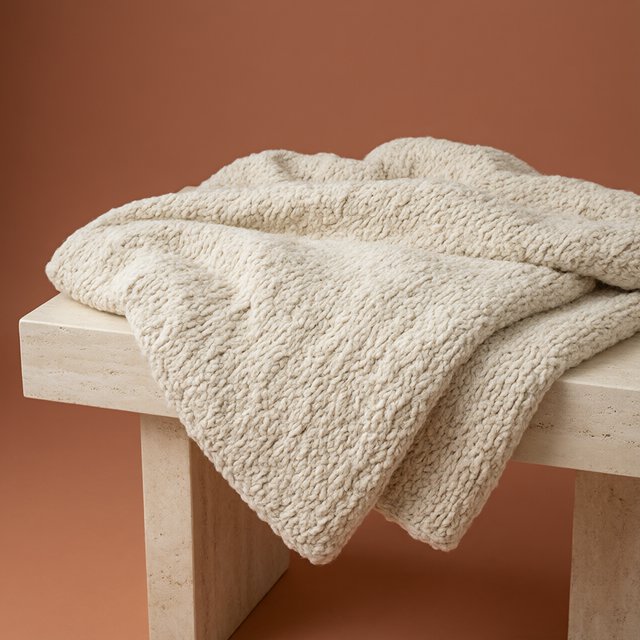 Soft folds of an unbranded chunky-knit wool throw rest on a pale travertine bench; its looped surface catches broad diffused key light and gentle side fill against a muted terracotta studio background, forming a clean commercial product image with no lettering. — Model 17
