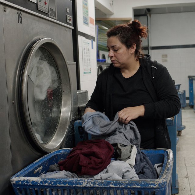 Candid documentary photograph of an adult woman sorting clothes in a self-service laundromat, cool fluorescent light, 35mm lens with moderate depth of field, loose lint suspended above a tumbling machine, worn baskets and tiled walls rendered naturally. — GPT Image 2