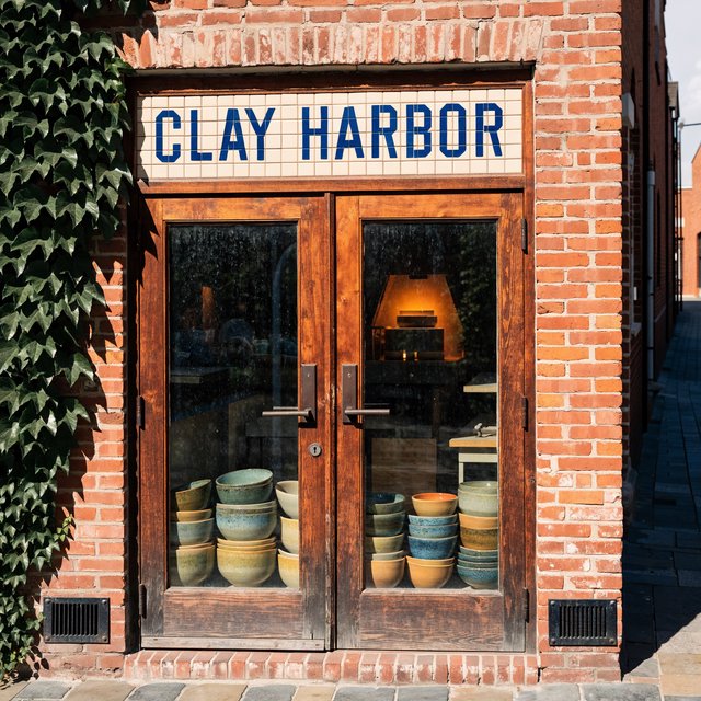 A sunlit ceramicist's studio storefront displays glazed bowls behind dusty glass doors; "CLAY HARBOR" is printed in cobalt-blue block letters across a cream tile transom above the entrance, with kiln warmth glowing inside. — Recraft v4 Pro