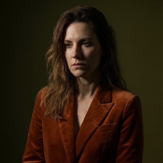 Her guarded gaze catches a solitary beam in a low-key fashion portrait: an adult woman aged 27–35 wears a rust-colored velvet blazer, the rest of her face falling into shadow against a deep olive backdrop. — Qwen Image 3.0 Pro