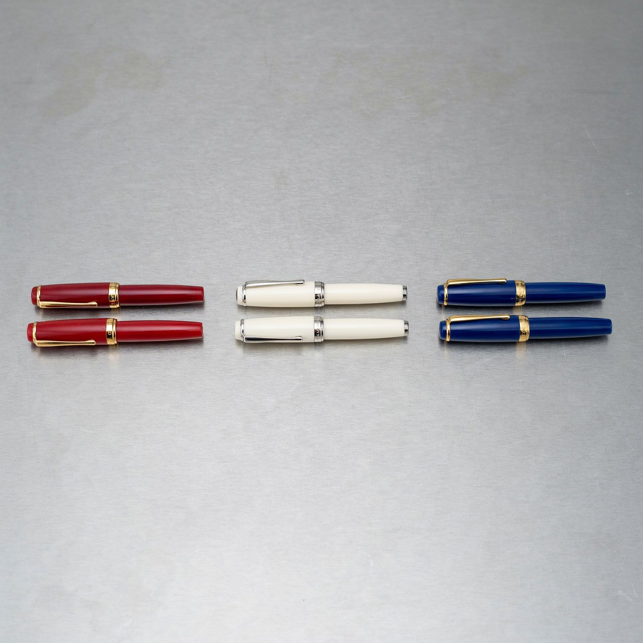 Across a brushed steel counter, exactly six fountain pens rest in three groups: two crimson pens on the left, two ivory pens in the center, and two cobalt pens on the right; nothing else. — Ideogram v4.5