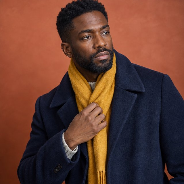 Editorial portrait of an adult Black man in his 30s wearing a deep navy wool overcoat and mustard scarf, fingers adjusting the scarf, thoughtful expression, muted terracotta studio backdrop, crisp cool window light. — GPT Image 2.5 Flare
