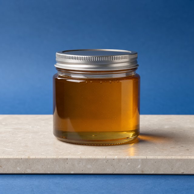 A squat unbranded honey jar with clear glass walls and a plain brushed-aluminum lid rests on honed pale limestone; broad diffused key light and warm rim light shape the commercial product image against a seamless cobalt-blue background, with no lettering. — Model 1