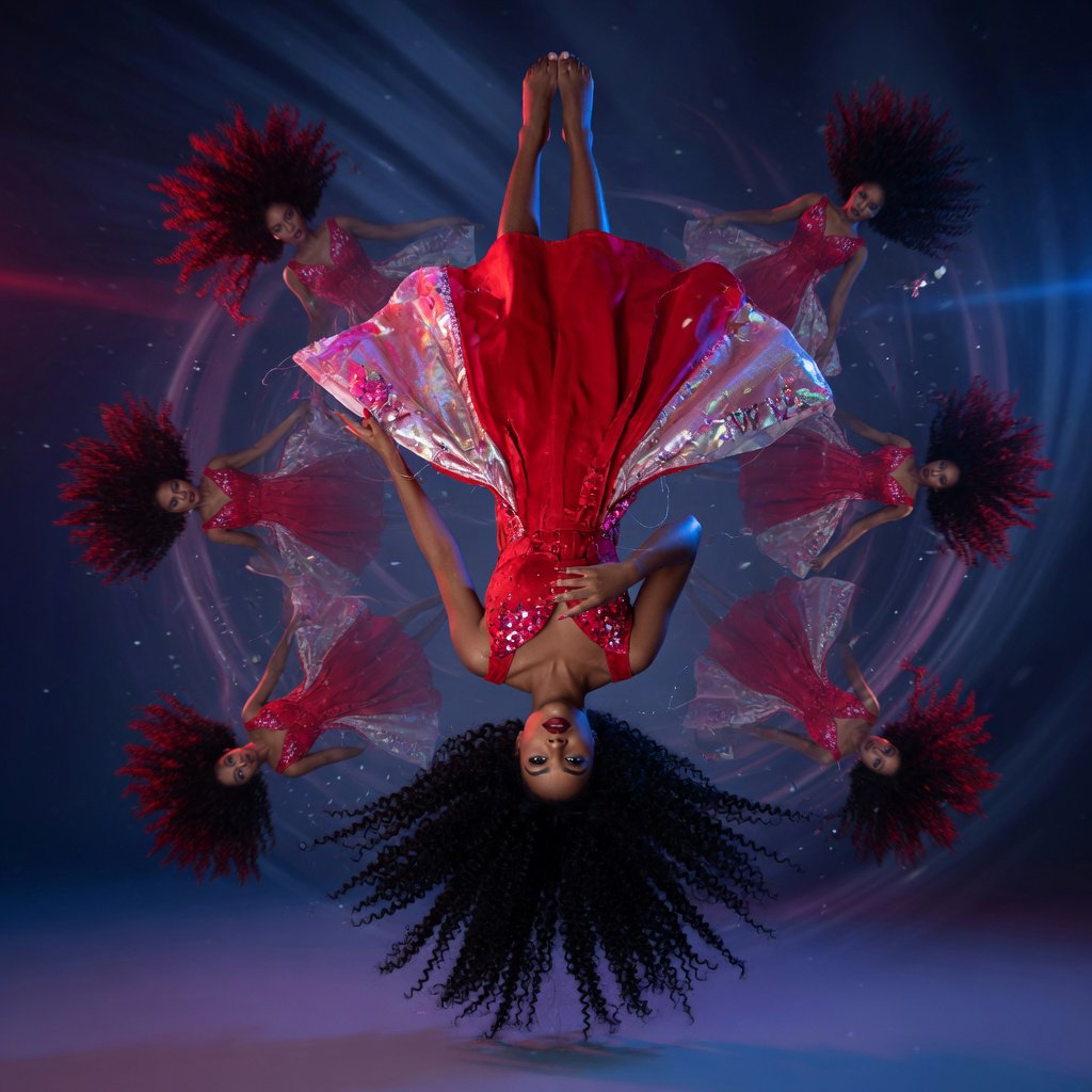 Image of a pop-dance African-American diva. The woman hangs completely inverted in mid-air, head pointing toward the floor and legs toward the ceiling. Her huge hair falls and spreads dramatically in the opposite direction, while her expression remains cool and composed. Her red gown is turned inside out, exposing its sparkling lining, seams, and construction. To push the surrealism further, a second, reversed version of her silhouette appears to peel outward from her original form, like a three-dimensional reflection escaping its owner. Multiple copies of the woman circle around a central point, each rotated to a different angle. Their flowing hair and red gowns form a pinwheel or circular vortex, creating the impression that one woman is spinning endlessly around herself. — Grok Imagine Image Quality