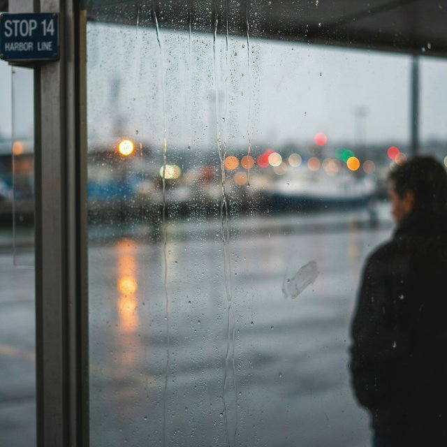 Raindrops crawl down a bus shelter pane, warping blurred harbor lights beyond. A handheld evening street snapshot in cool sodium glow, shot at 50mm with shallow depth of field, catches tiny water trails and a faint wipe mark. — Ideogram v4