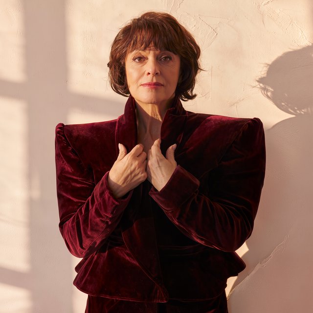 Fashion portrait of an adult woman in her early 50s wearing a sculptural burgundy velvet jacket, her hands lightly gathering its lapel. A wry, composed expression meets the camera against a warm ivory plaster backdrop, lit by soft late-afternoon window glow. — Kling v3