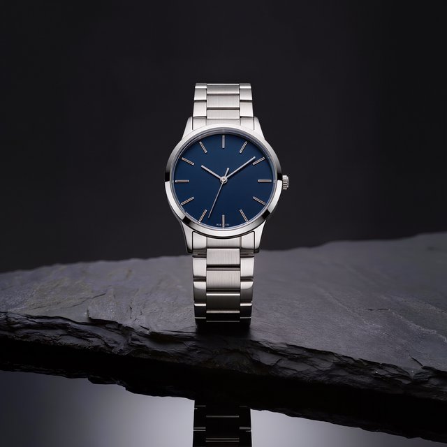Product shot of a minimalist steel wristwatch with a navy dial on dark slate, single softbox from the upper left, subtle reflection. — Kling v3
