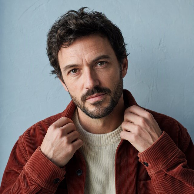Portrait photograph of an adult man in his late 30s wearing a rust-colored corduroy overshirt over a cream knit, fingertips resting on its broad collar. His quietly curious gaze meets the camera against a pale blue plaster backdrop, softly lit by cool side-window light. — GPT Image 2