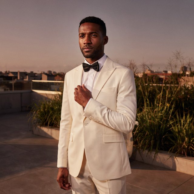Under the copper glow of a rooftop terrace, an adult Black man in a tailored cream dinner suit is caught smoothing his lapel as evening breeze stirs nearby grasses; calm poise, sculpted shadows and refined fashion-magazine polish. — Ideogram v4.5