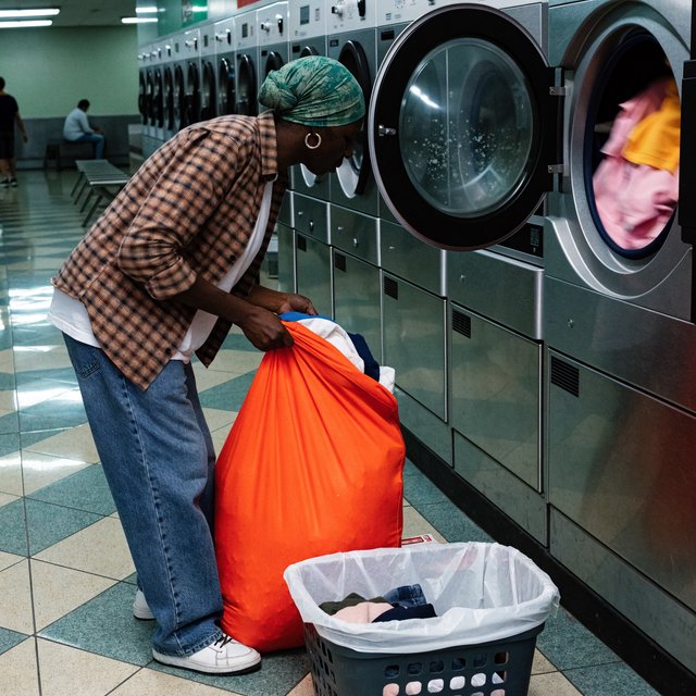 Candid documentary photograph of an adult sorting laundry in a neighborhood laundromat, cool fluorescent light, 35mm lens with moderate depth of field, lint specks floating near a tumbling dryer door. — Recraft v4 Pro