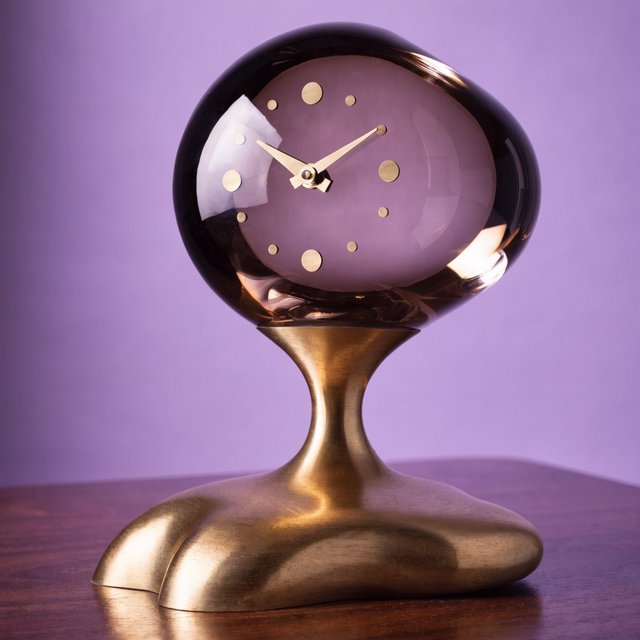 Commercial product photograph of an unbranded sculptural table clock made from satin brass and smoked glass, resting on a dark walnut surface against a muted lavender background, illuminated by a large diffused softbox from the upper left and a subtle rear rim light. — Recraft v4