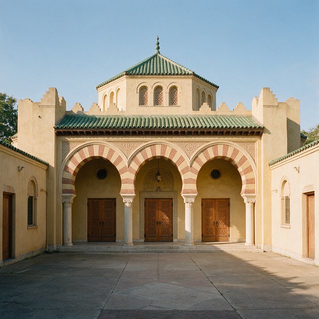 Exterior of a Moorish Revival municipal bathhouse, pale buff stucco, striped brick arches, green glazed tile roof and carved cedar doors; clear morning light, viewed across a paved courtyard, with coherent structure and crisp lines. — Seedream 5.0 Pro