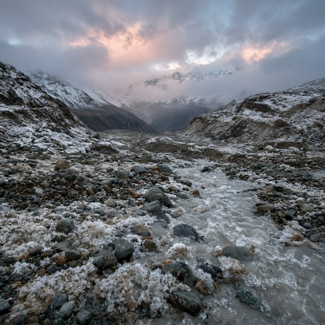Beyond a braided glacier-fed stream, a steep moraine valley opens beneath clearing dawn snow flurries; gritty ice grains and water-polished stones sharpen the near bank, while layered ridges fade into cloud. — Model 5