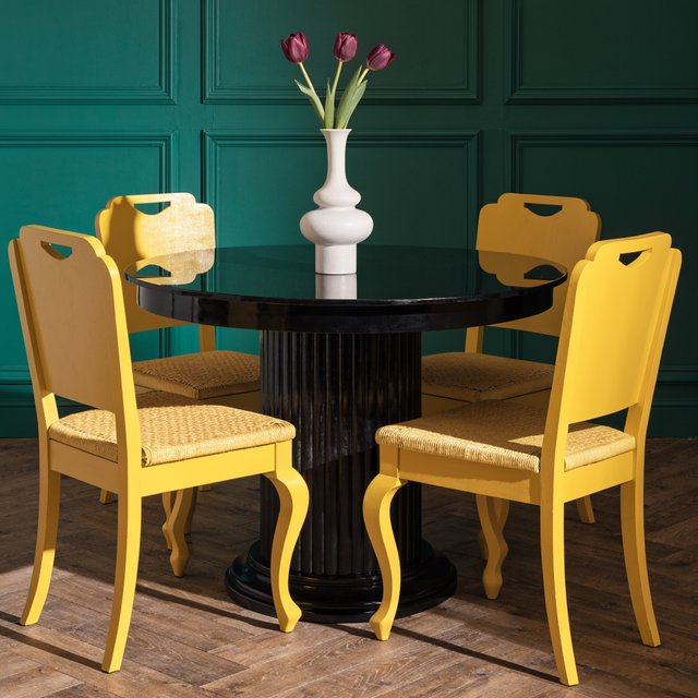 Exactly four yellow wooden chairs surround a round black table: two chairs at the front, one on the left, and one on the right. A white vase with three purple tulips sits at the table center, nothing else. — Recraft v4