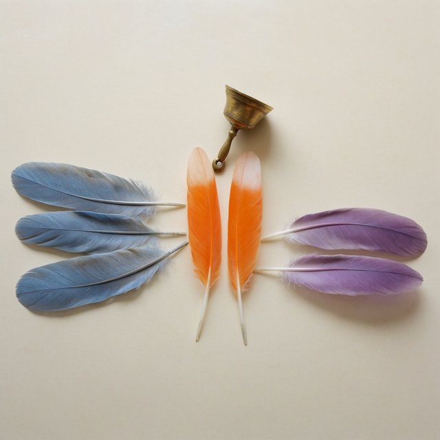 Exactly seven feathers lie on a cream tabletop: three blue on the left, two orange in the center, and two purple on the right. A brass bell sits above the orange pair, nothing else. — Krea 2 Large