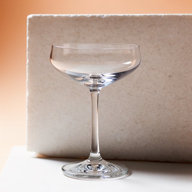 Beside a pale limestone block, an unbranded handblown cocktail coupe in clear soda-lime glass catches crisp window-shaped highlights; its curved bowl bends a muted apricot backdrop under broad diffused key light and narrow cool rim lighting, commercial product image, no lettering. — Krea 2 Large