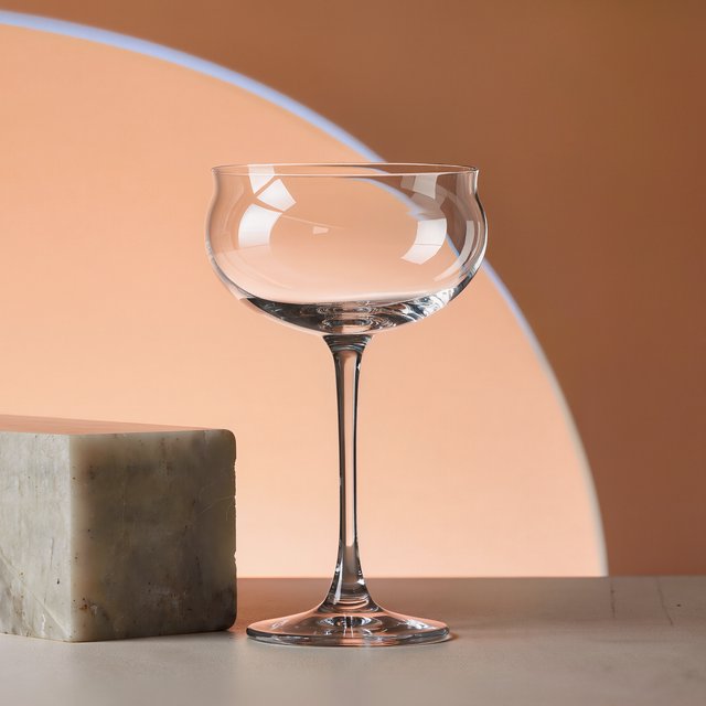 Beside a pale limestone block, an unbranded handblown cocktail coupe in clear soda-lime glass catches crisp window-shaped highlights; its curved bowl bends a muted apricot backdrop under broad diffused key light and narrow cool rim lighting, commercial product image, no lettering. — Kling v3