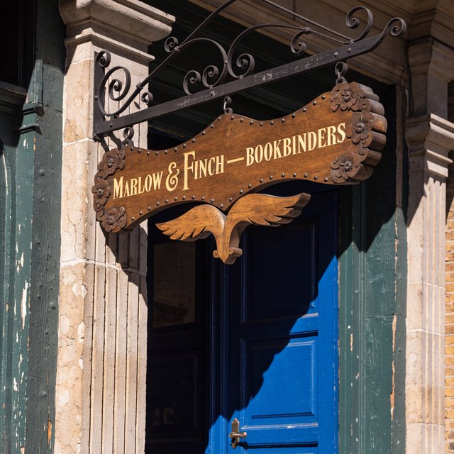 A hanging wooden shop sign that reads "MARLOW & FINCH — BOOKBINDERS" in gold serif letters, carved, above a blue door. — Recraft v4