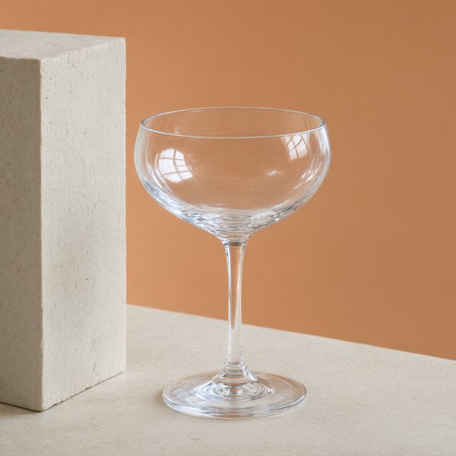 Beside a pale limestone block, an unbranded handblown cocktail coupe in clear soda-lime glass catches crisp window-shaped highlights; its curved bowl bends a muted apricot backdrop under broad diffused key light and narrow cool rim lighting, commercial product image, no lettering. — Qwen Image 3.0