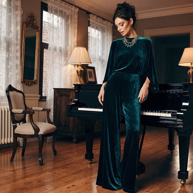 Editorial glamour photograph of an adult woman in a midnight-green velvet evening jumpsuit, standing naturally beside a grand piano in a softly lit salon, warm flattering light, relaxed hands, elegant posture, sophisticated fashion composition. — Recraft v4 Pro