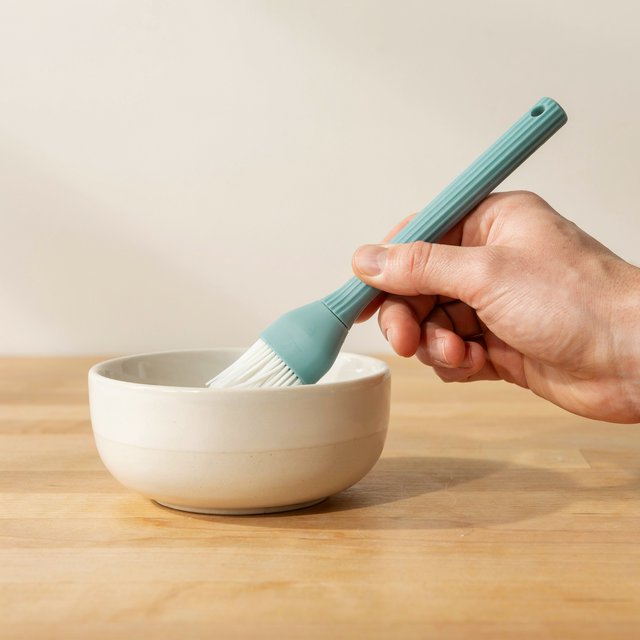 Across a honed maple counter, an adult hand guides an unbranded silicone pastry brush over a ceramic bowl; its ribbed teal handle catches diffused key light and warm bounce against a cream kitchen background, face cropped out, no lettering, commercial product image. — Ideogram v4.5