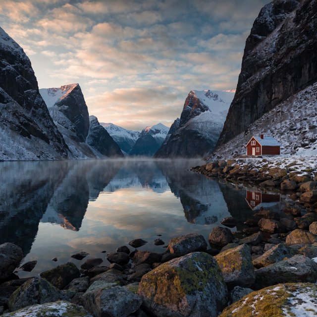 A Norwegian fjord at dawn, mist over still water, a single red cabin on the shore, snow on the peaks, wide angle. — Recraft v4
