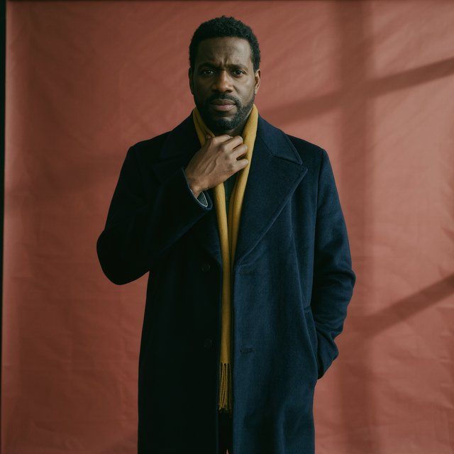 Editorial portrait of an adult Black man in his 30s wearing a deep navy wool overcoat and mustard scarf, fingers adjusting the scarf, thoughtful expression, muted terracotta studio backdrop, crisp cool window light. — Kling v3 Omni