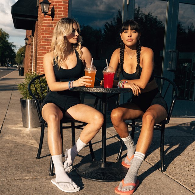 Two muscular adult women in their twenties, one Caucasian blonde and one Latina, sit at an outdoor café table beside a gym after their workout. They wear tight workout shorts, sports bras, and flip-flops. — Recraft v4