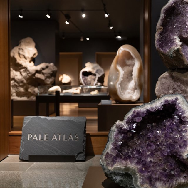 Within a quiet mineral gallery, amethyst clusters and polished geodes catch pools of light; "PALE ATLAS" is carved in clean capitals across a slate plaque beside the entrance. — ImagineArt 2