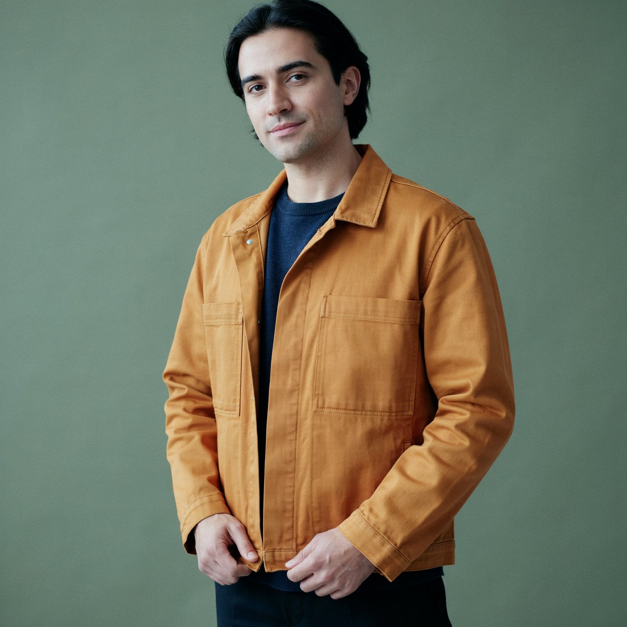 Fashion portrait of an adult nonbinary person in their late twenties wearing a structured saffron-yellow canvas jacket over a fine-gauge navy merino top, hands resting on the jacket hem. Their bright, self-assured expression contrasts with a softly lit sage-green backdrop. — Ideogram v4