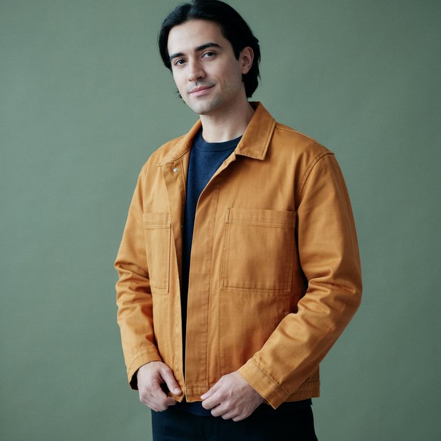 Fashion portrait of an adult nonbinary person in their late twenties wearing a structured saffron-yellow canvas jacket over a fine-gauge navy merino top, hands resting on the jacket hem. Their bright, self-assured expression contrasts with a softly lit sage-green backdrop. — Ideogram v4