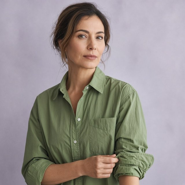 Portrait photograph of an adult woman in her late 30s wearing a moss-green cotton poplin shirt, fingers lightly turning its rolled cuff. Her calm, observant expression faces the camera against a chalky lavender backdrop, lit by soft diffused skylight. — Grok Imagine Image 2