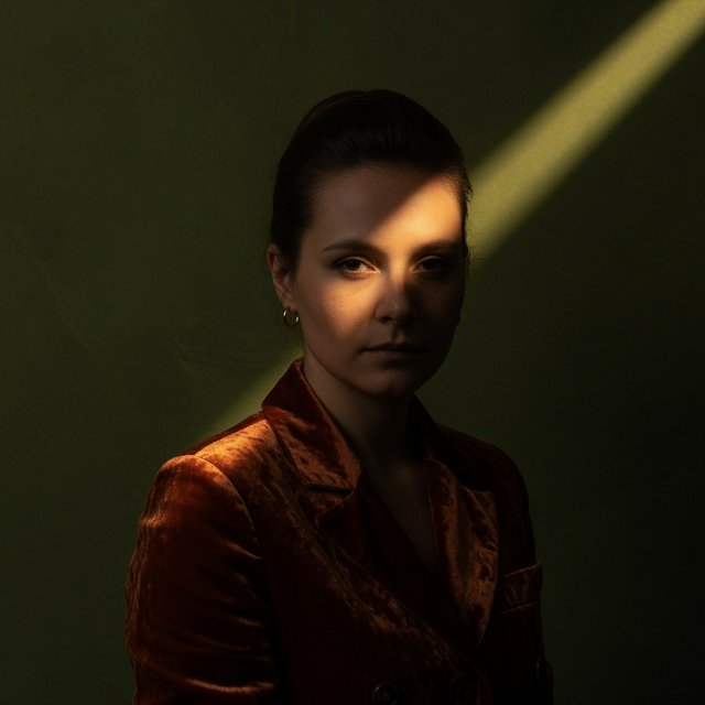 Her guarded gaze catches a solitary beam in a low-key fashion portrait: an adult woman aged 27–35 wears a rust-colored velvet blazer, the rest of her face falling into shadow against a deep olive backdrop. — Grok Imagine Image Quality