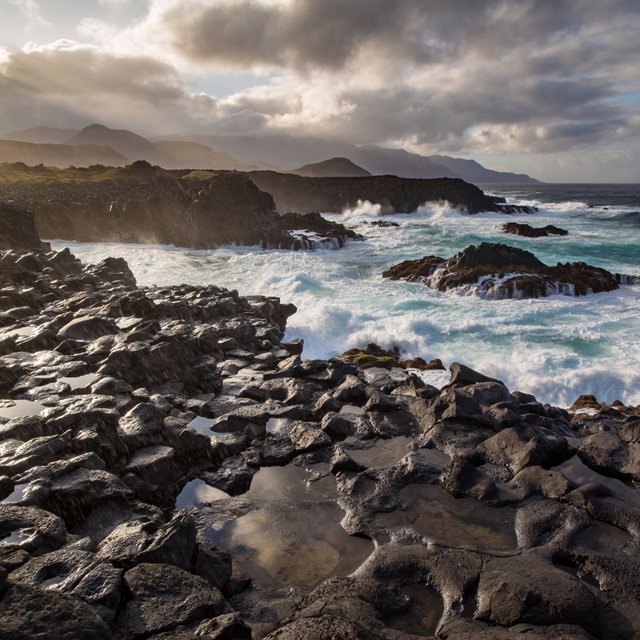 A rugged volcanic coastline under clearing storm clouds at late afternoon, black basalt shelves descending into turquoise surf, sea spray clinging to jagged rocks, tide pools reflecting distant cliffs and layered mountains fading into rain haze. — ImagineArt 2