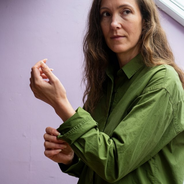 Portrait photograph of an adult woman in her late 30s wearing a moss-green cotton poplin shirt, fingers lightly turning its rolled cuff. Her calm, observant expression faces the camera against a chalky lavender backdrop, lit by soft diffused skylight. — Krea 2 Large