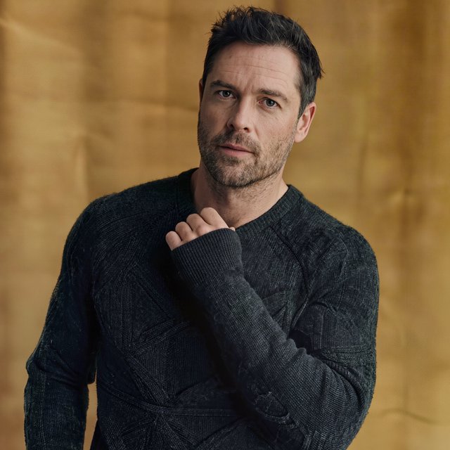 Editorial portrait of an adult man in his early 40s wearing a charcoal wool fisherman’s sweater, one hand loosely holding its ribbed cuff. His thoughtful, slightly tired expression rests against a muted ochre studio backdrop, shaped by gentle directional window light. — Kling v3
