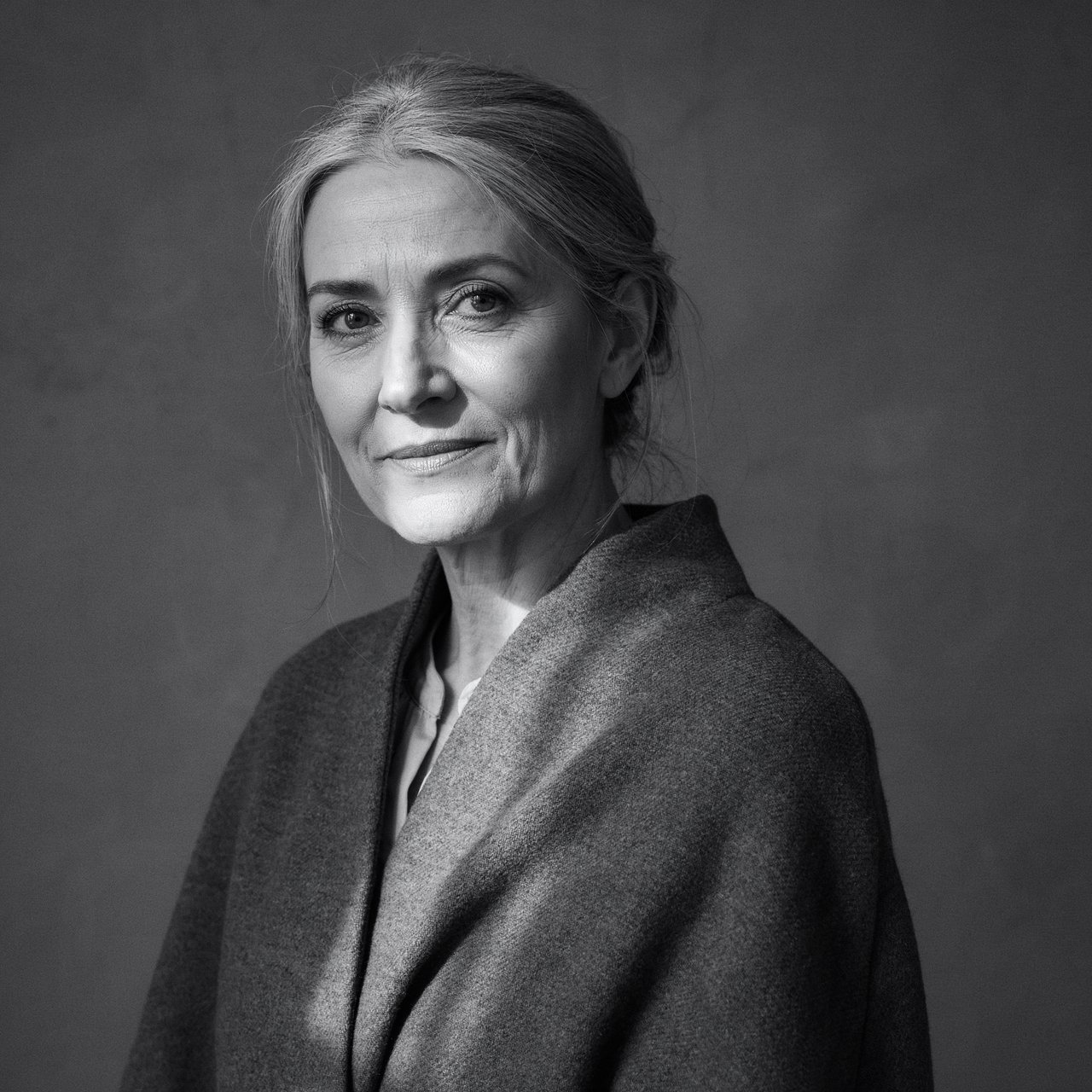 Under a narrow window beam, an adult woman in her late 50s wears a sculpted wool cape over a cotton blouse; her wry expression rests against a charcoal plaster backdrop in a black-and-white portrait, shaped by soft angled light. — Ideogram v4