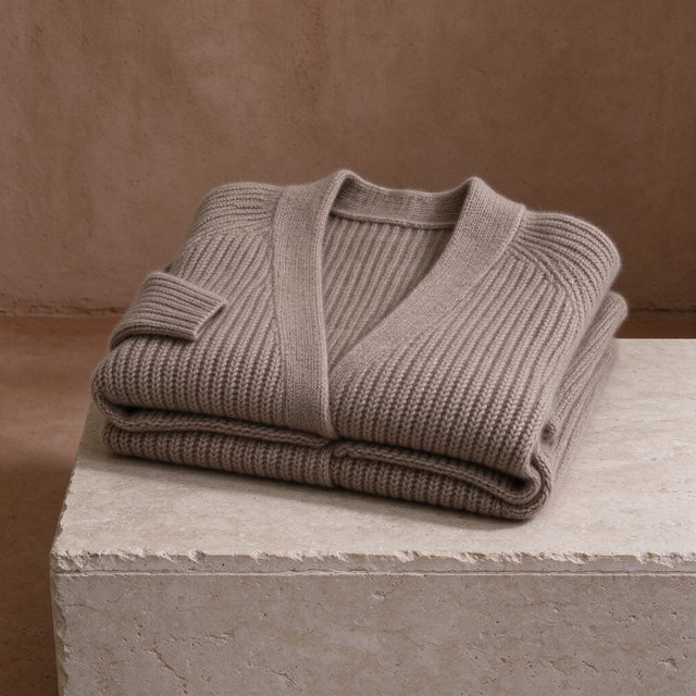 A folded unbranded merino cardigan rests on a honed limestone plinth, its chunky ribbed wool surface softly catching broad overhead diffusion and warm side bounce against a muted clay backdrop, with no tags, lettering, or graphics. — Seedream 5.0 Flash