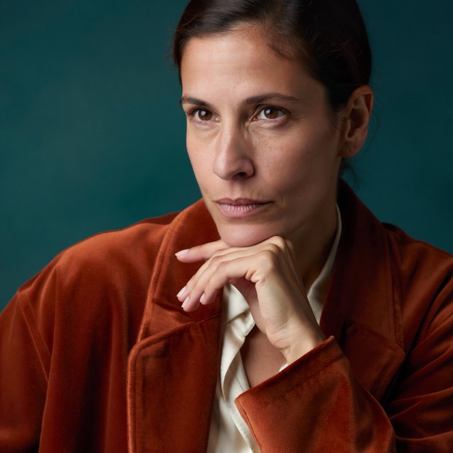 Portrait of an adult woman in her 30s wearing a rust-colored velvet jacket over an ivory blouse, one hand resting beneath her chin, thoughtful expression, deep teal studio backdrop, soft side lighting and gentle cheek highlights. — ImagineArt 2
